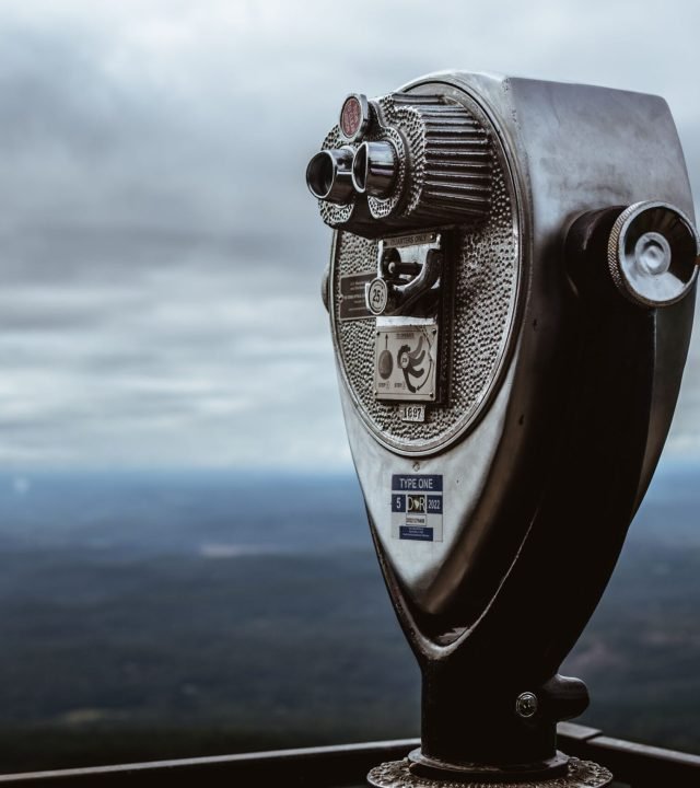 A metal coin-operated binocular telescope on a mountain with a cloudy sky in the background.