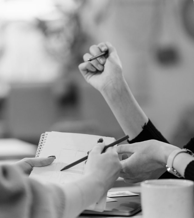 Black and white image of people collaborating at a desk, focusing on hands writing.